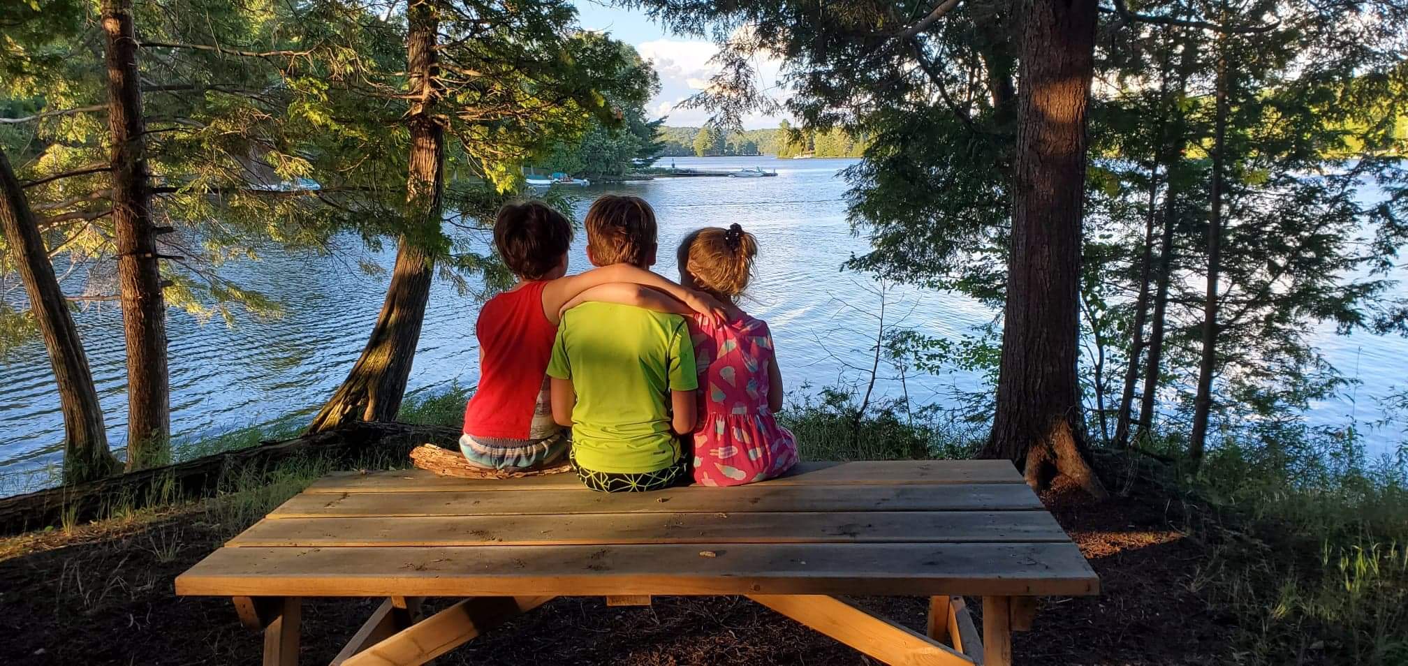 three kids, sitting on picnic table looking out over a lake