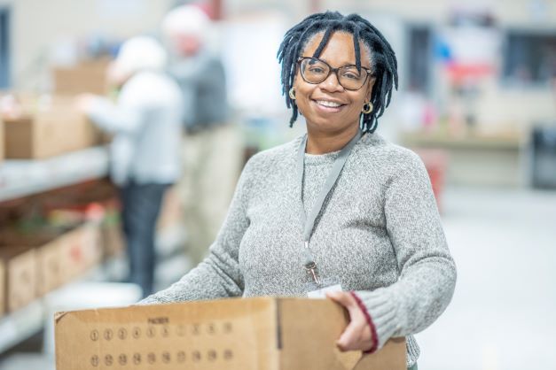 woman holding a cardboard box, smiling as she volunteers at food program